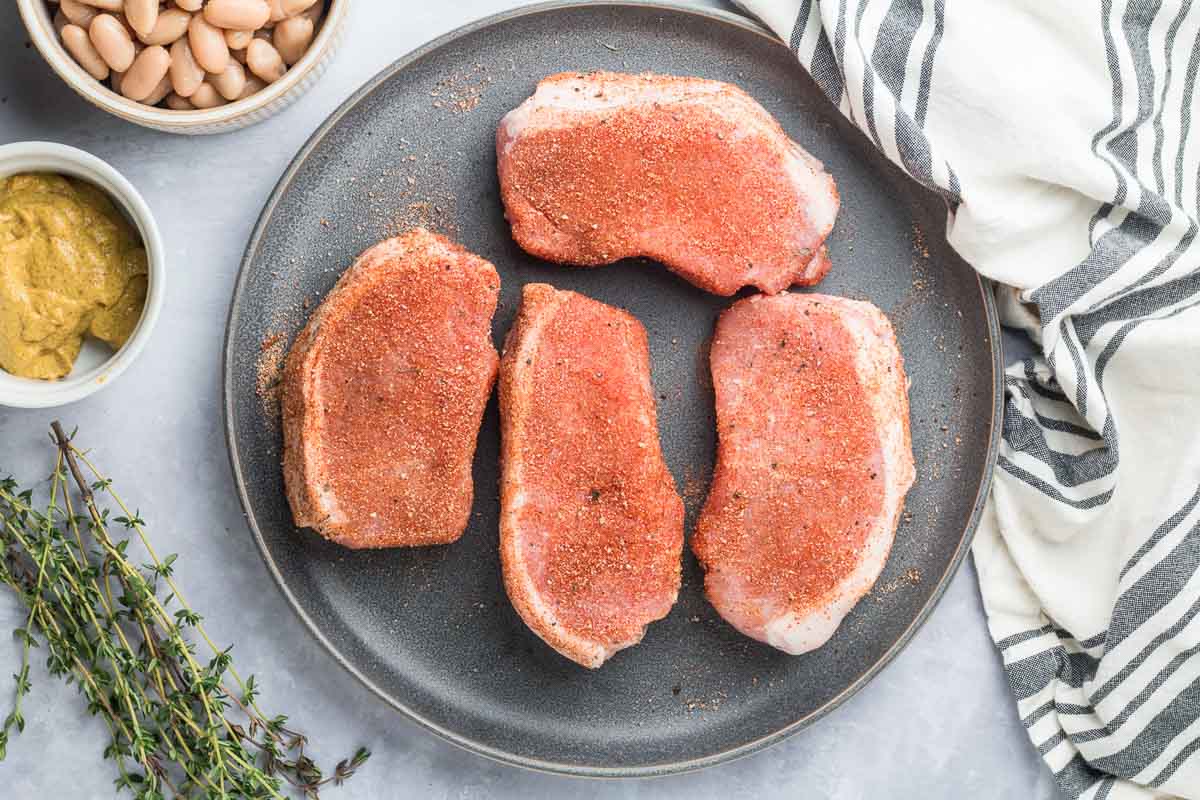 Four raw pork chops coated with dry seasoning sit on a dark plate-perfect for a quick and easy Skillet Pork Chops dinner on busy school nights. Nearby are a striped towel, fresh thyme, a bowl of mustard, and a bowl of beans.