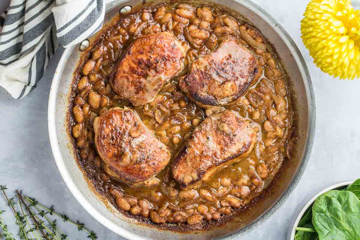 Skillet Pork Chops are paired with seasoned white beans in a rich brown sauce, making this the perfect meal for busy School Nights. Four chops sit in the skillet next to a cloth napkin, yellow flower, and fresh spinach leaves.