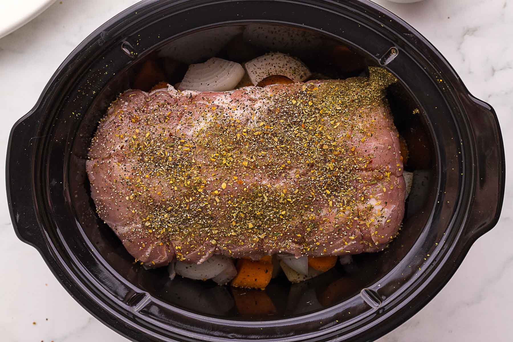 Uncooked seasoned pork roast placed on top of chopped vegetables inside a black slow cooker on a white surface.