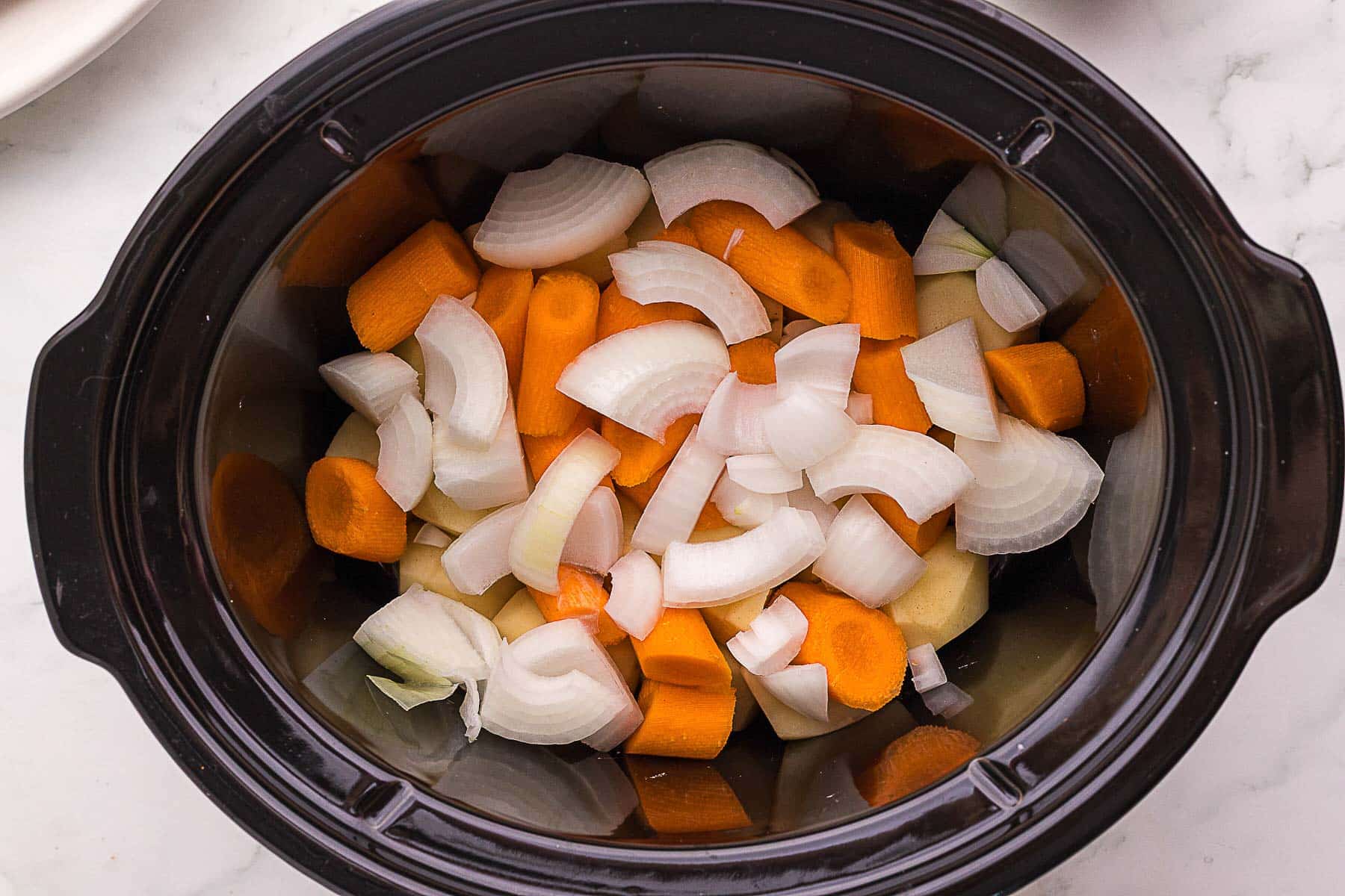 Chopped onions, carrots, and potatoes are placed in a black slow cooker on a light-colored surface.