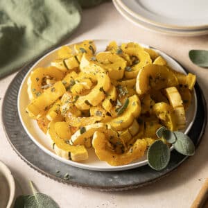 A plate of roasted delicata squash slices garnished with chopped herbs, set on a table with plates and sage leaves nearby.