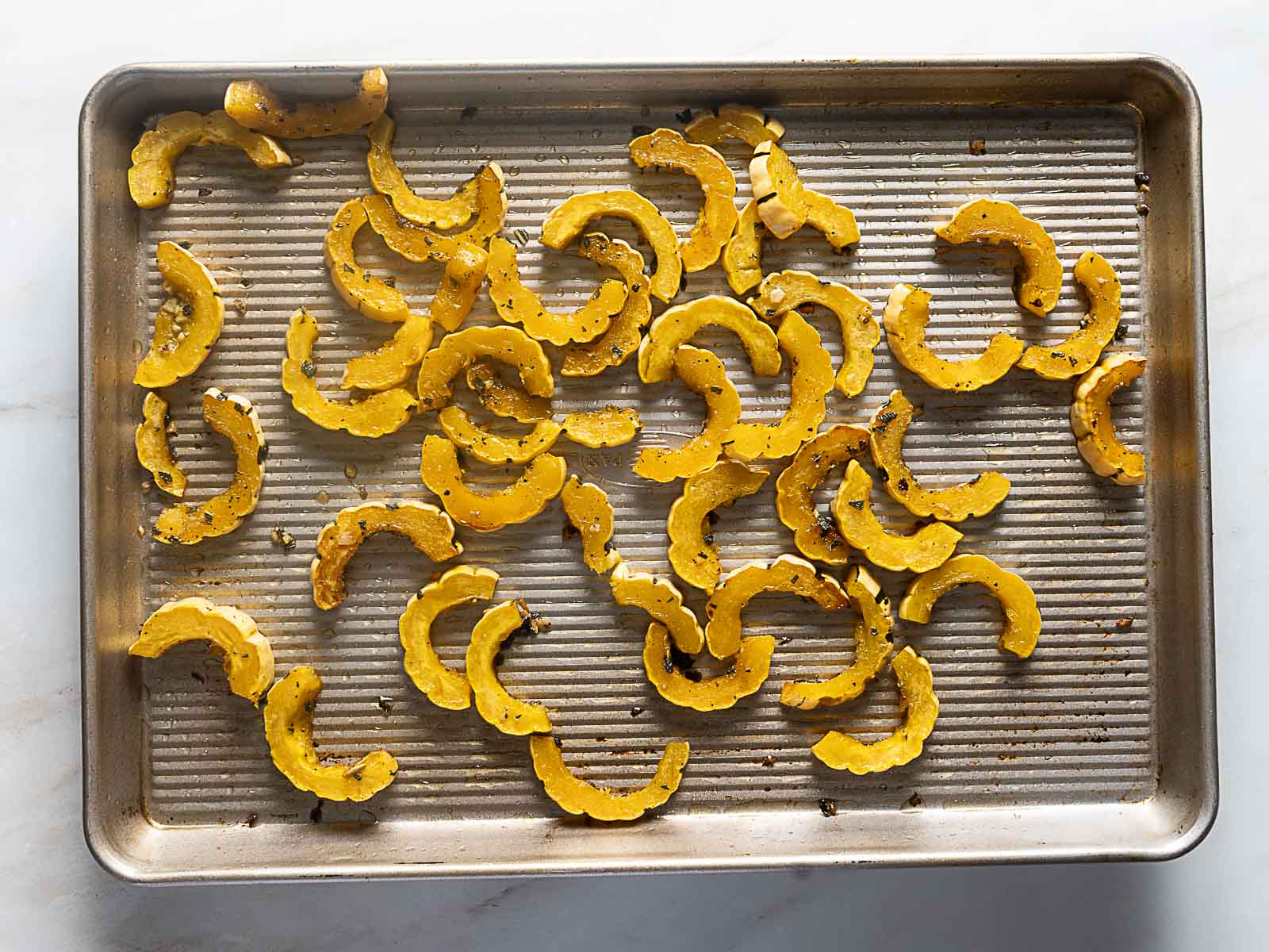 Sliced delicata squash pieces spread out on a metal baking sheet, seasoned and ready to be roasted.