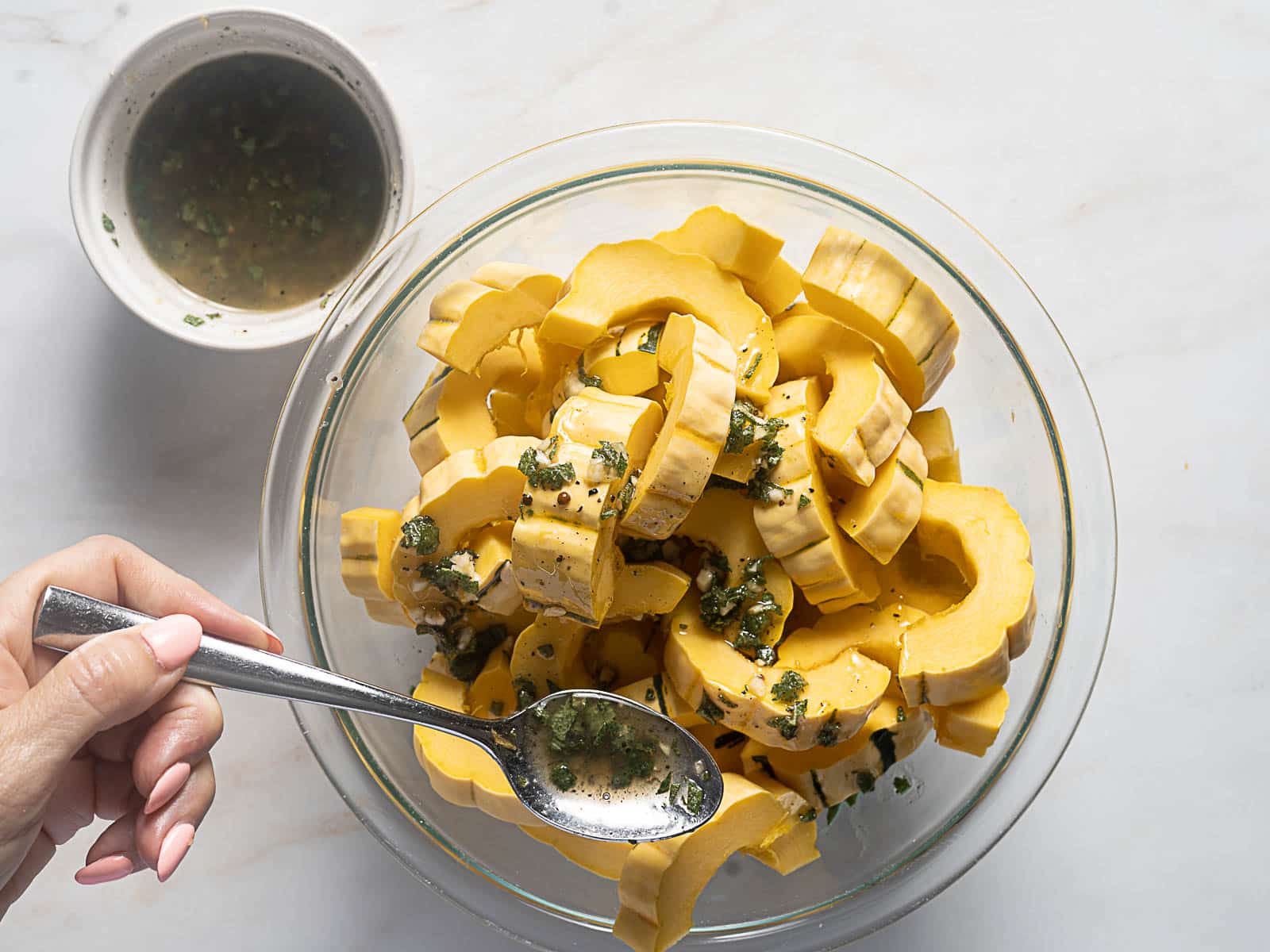 A hand spoons herb dressing over sliced delicata squash in a glass bowl, with extra dressing in a small bowl nearby on a white surface.