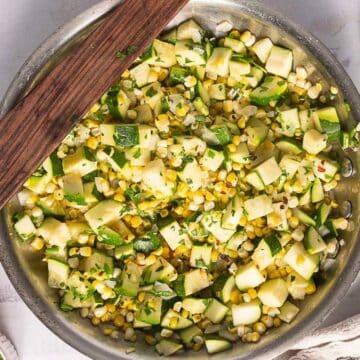 A skillet filled with sautéed diced zucchini, corn, and herbs sits on a light-colored surface with a wooden spatula resting on top—a perfect addition to your collection of corn recipes to warm the soul.
