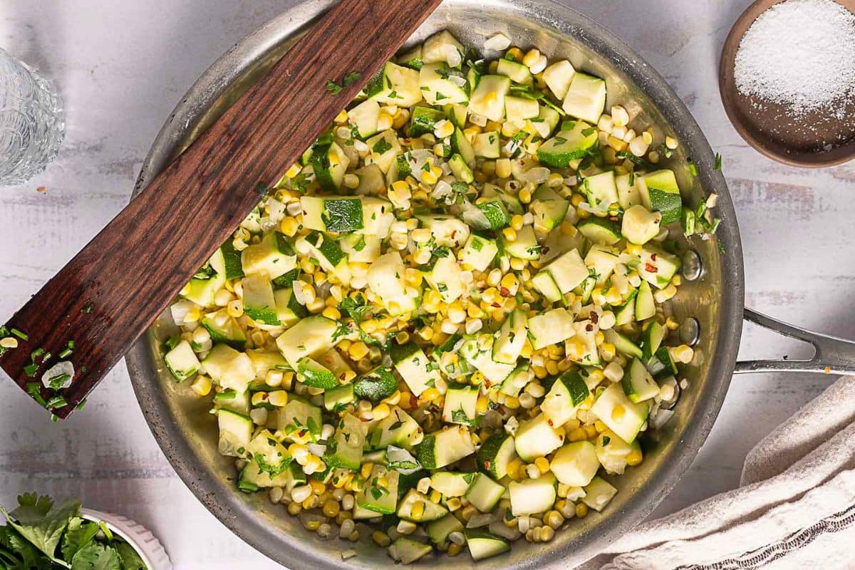 A skillet filled with sautéed diced zucchini, corn, and herbs sits on a light-colored surface with a wooden spatula resting on top—a perfect addition to your collection of corn recipes to warm the soul.
