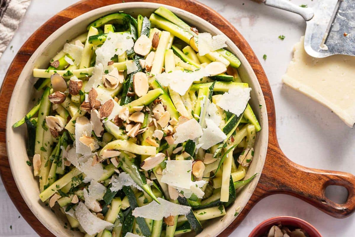 A wooden bowl of zucchini salad topped with sliced almonds and cheese shavings, placed on a round wooden serving board-perfect for easy side dishes at weeknight sides or dinner sides gatherings.