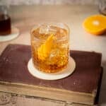 A glass of amber cocktail with ice and an orange twist sits on a coaster atop a closed vintage book, with half an orange and another drink in the background.