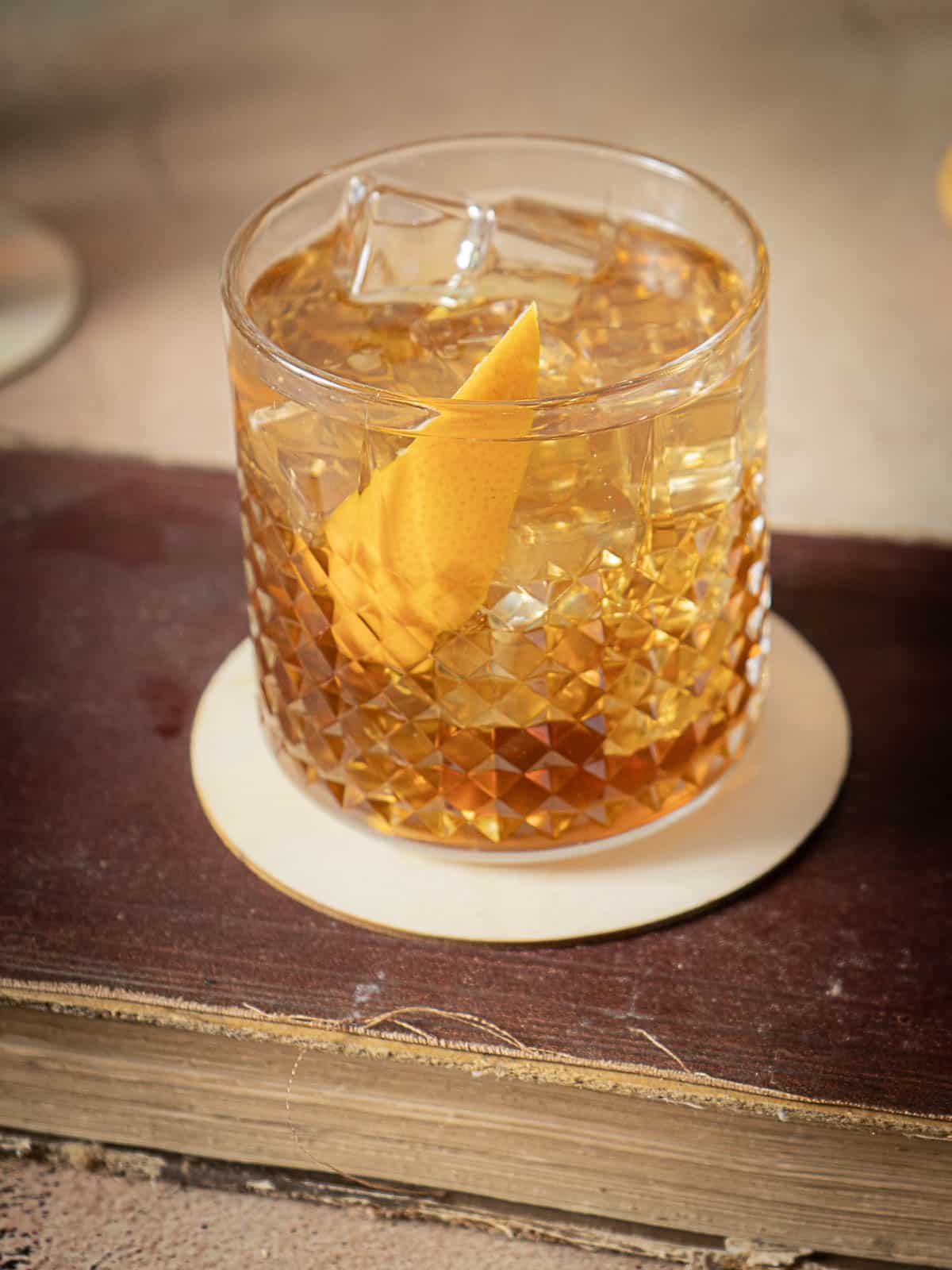 A glass of amber-colored cocktail with ice and an orange peel garnish, placed on a coaster atop a closed, old book.