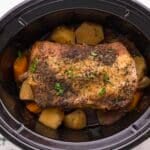 Seasoned pork roast sits atop chopped potatoes and carrots inside a black slow cooker.
