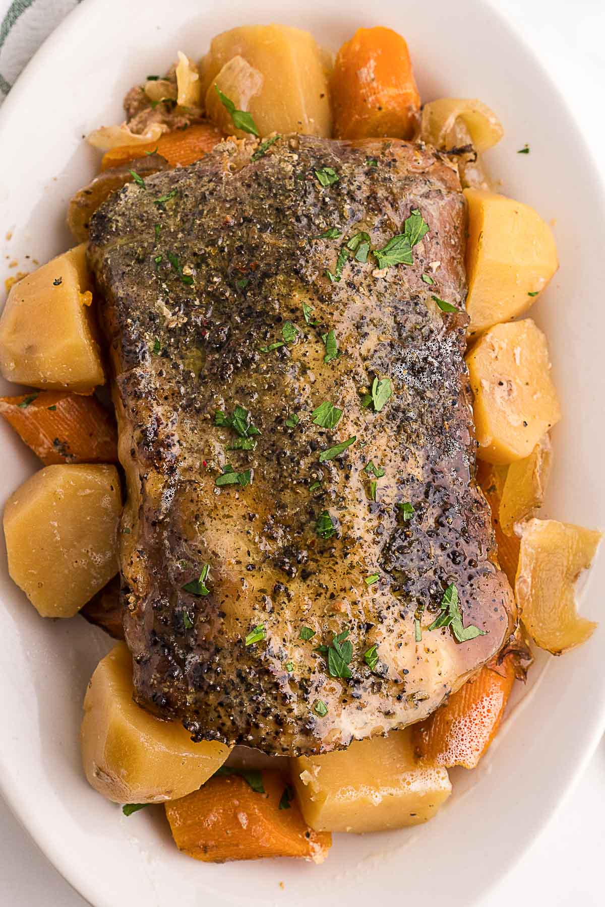 Seasoned roast pork on a white platter, surrounded by chunks of cooked potatoes and carrots, garnished with chopped herbs.