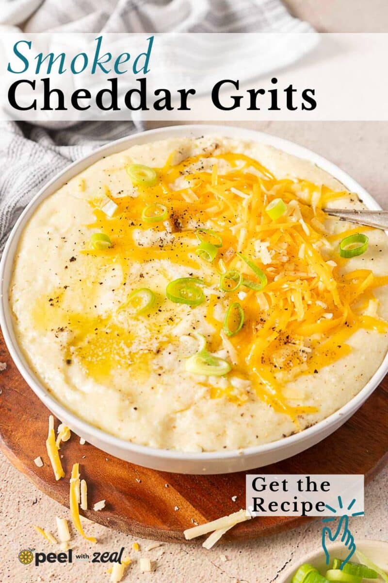 A bowl of creamy cheddar grits topped with shredded cheddar cheese, sliced green onions, and cracked black pepper, set on a wooden surface with a spoon.