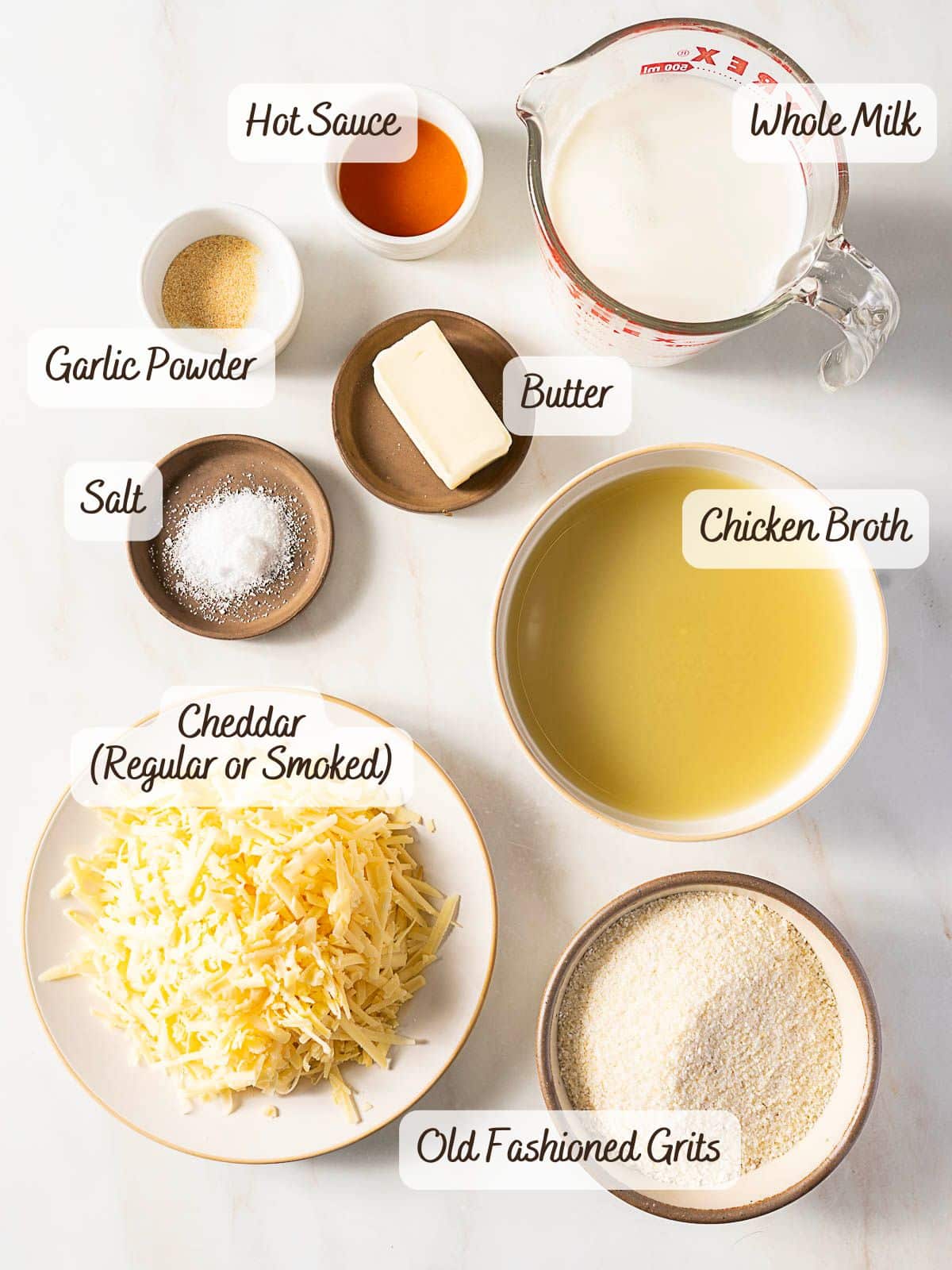 Ingredients for cheddar grits are arranged on a counter, including whole milk, chicken broth, hot sauce, butter, garlic powder, salt, cheddar cheese, and old fashioned grits.