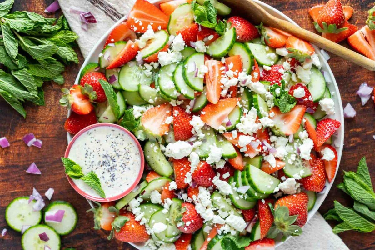 A salad with sliced strawberries, cucumbers, red onions, crumbled feta, and fresh mint is served on a plate with a small bowl of creamy dressing-one of the best easy side dishes for weeknight recipes.