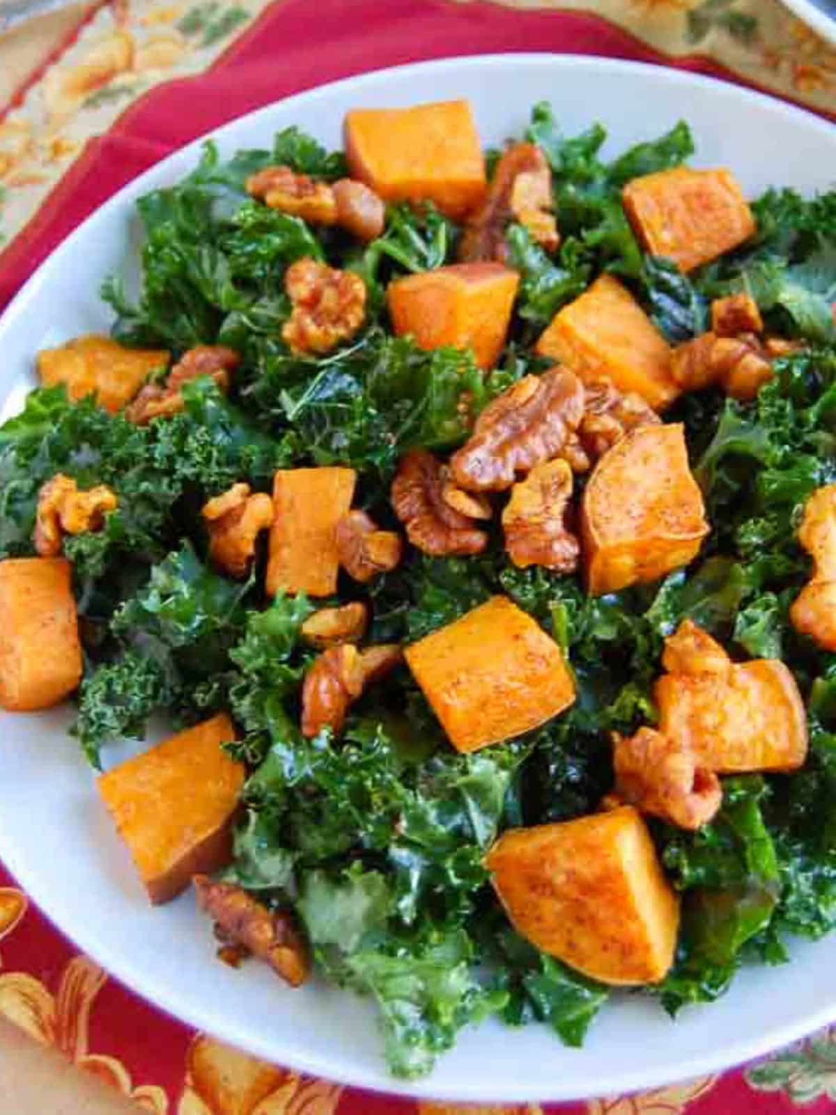 A salad with kale, roasted sweet potato cubes, and walnuts served on a white plate.