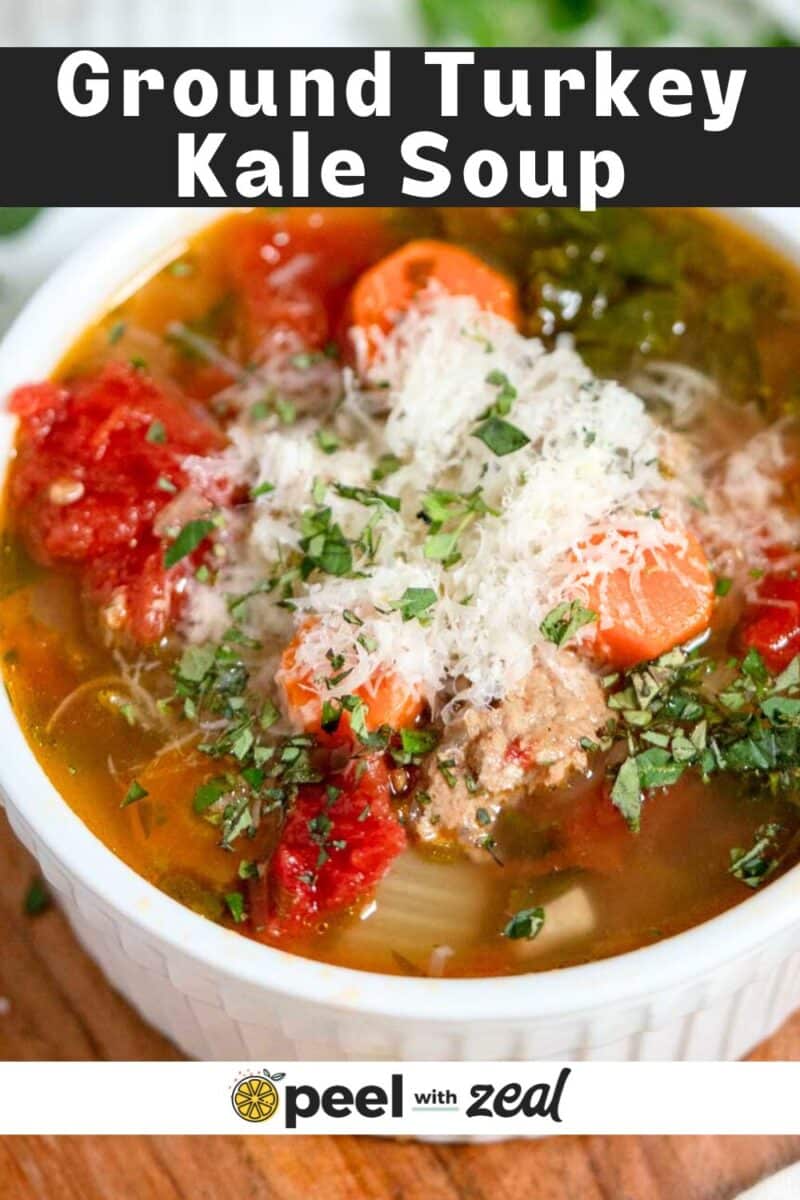 A bowl of ground turkey kale soup with carrots, tomatoes, shredded cheese, and herbs, shown in a white bowl on a wooden surface.