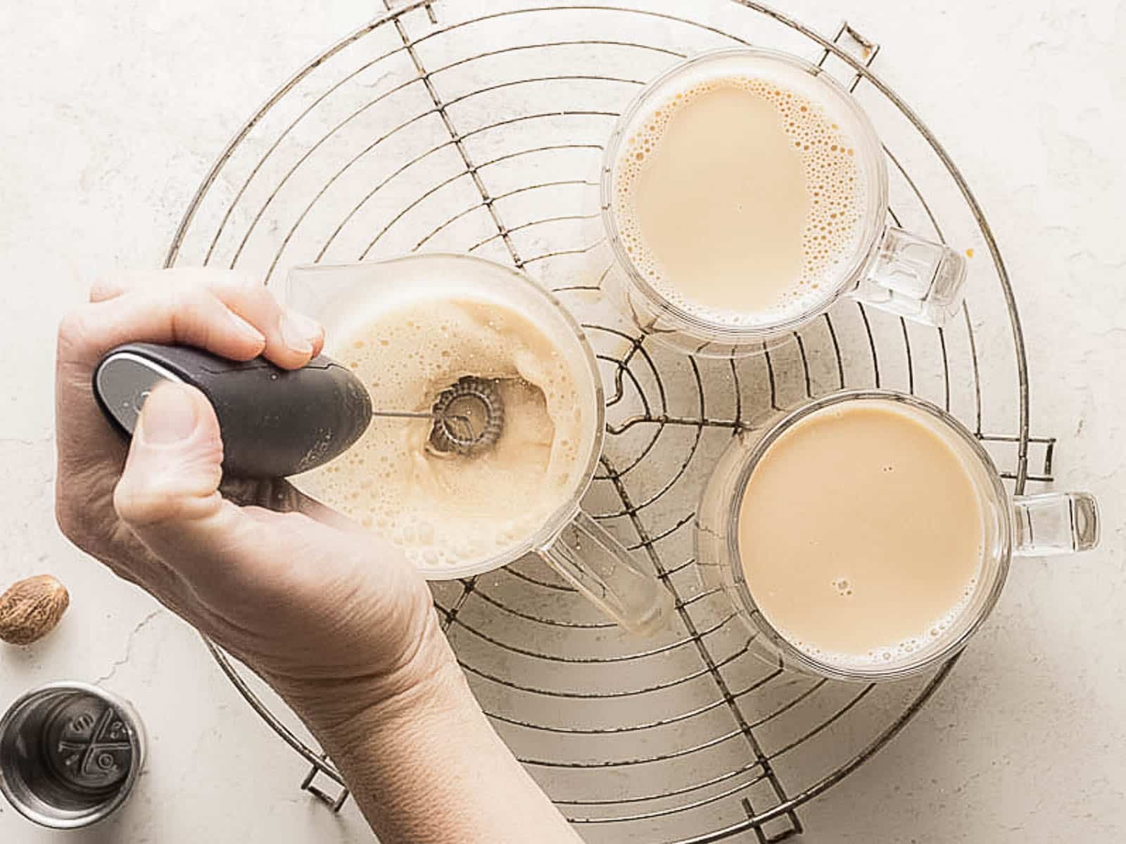 A hand uses a milk frother in a glass mug filled with milk tea, next to two other glass mugs of milk tea on a round metal cooling rack.