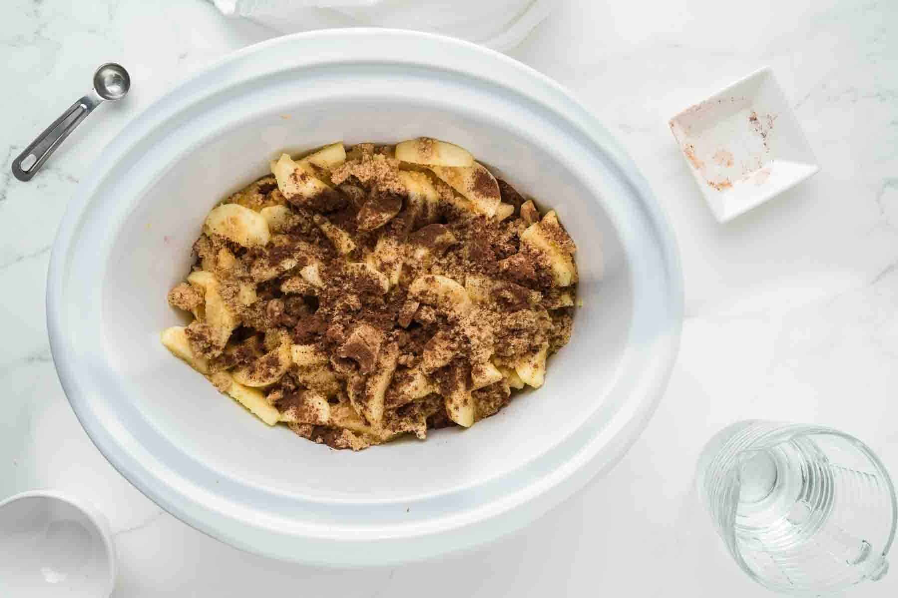 Sliced apples topped with brown sugar and cinnamon in a white slow cooker, with measuring spoon, glass, and bowl on a white countertop.