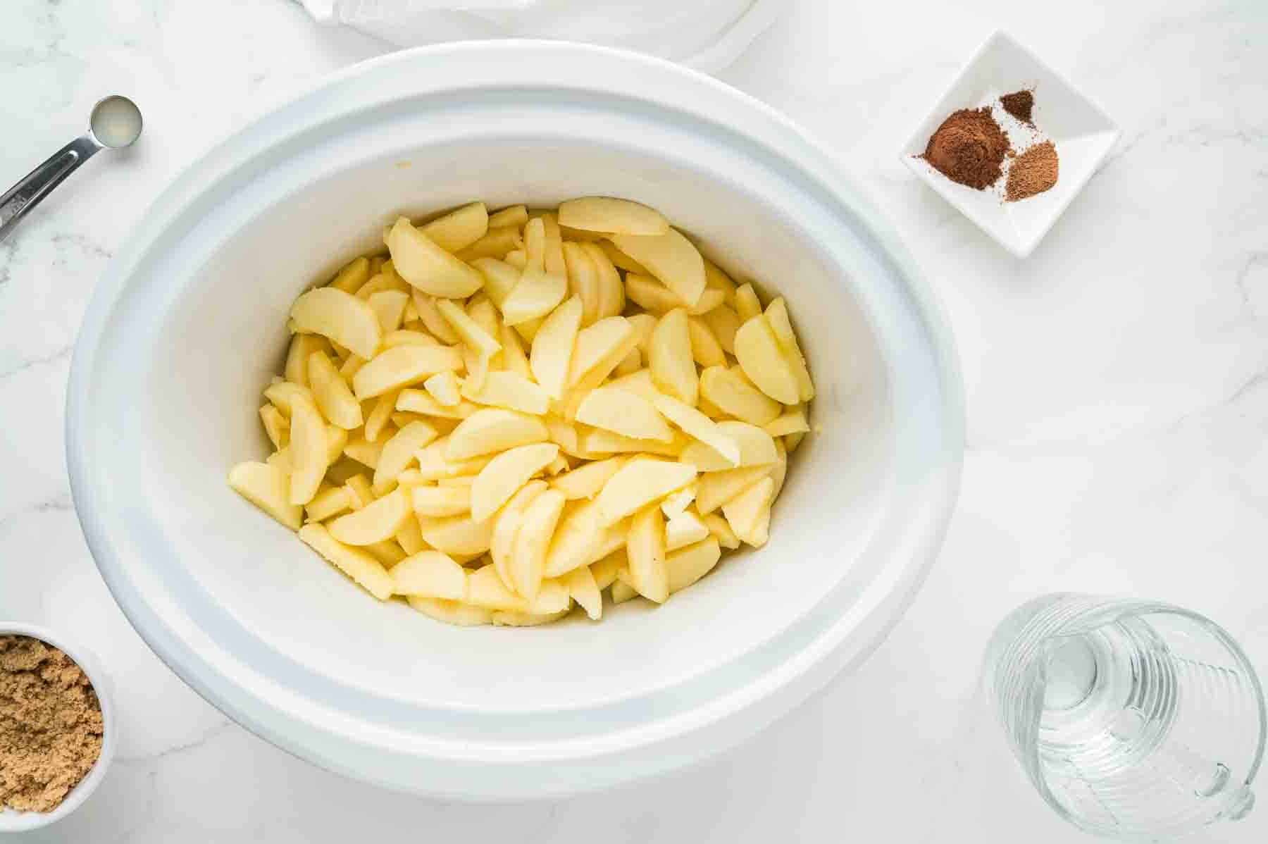 Sliced apples in a white slow cooker, with bowls of brown sugar, spices, and a glass of water on a white countertop.