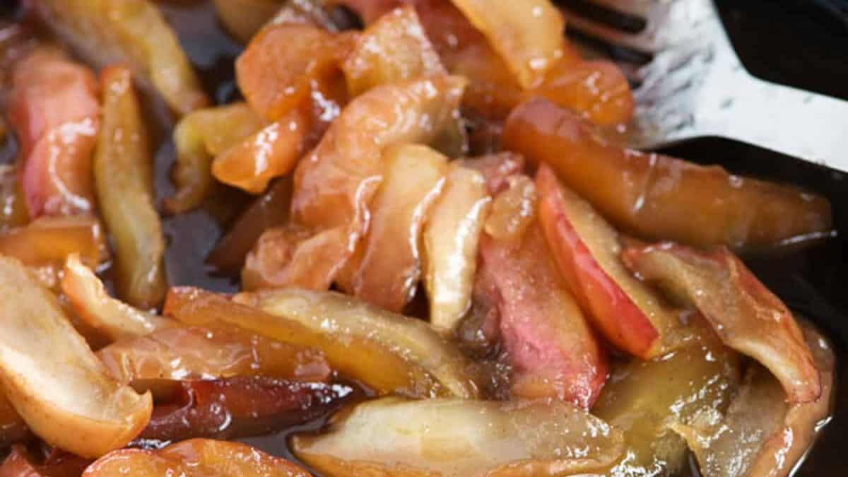 Close-up of caramelized apple slices in a pan, glistening with syrup and partially cooked, with kitchen utensils visible in the background—a delicious inspiration for seasonal recipes and apple desserts.