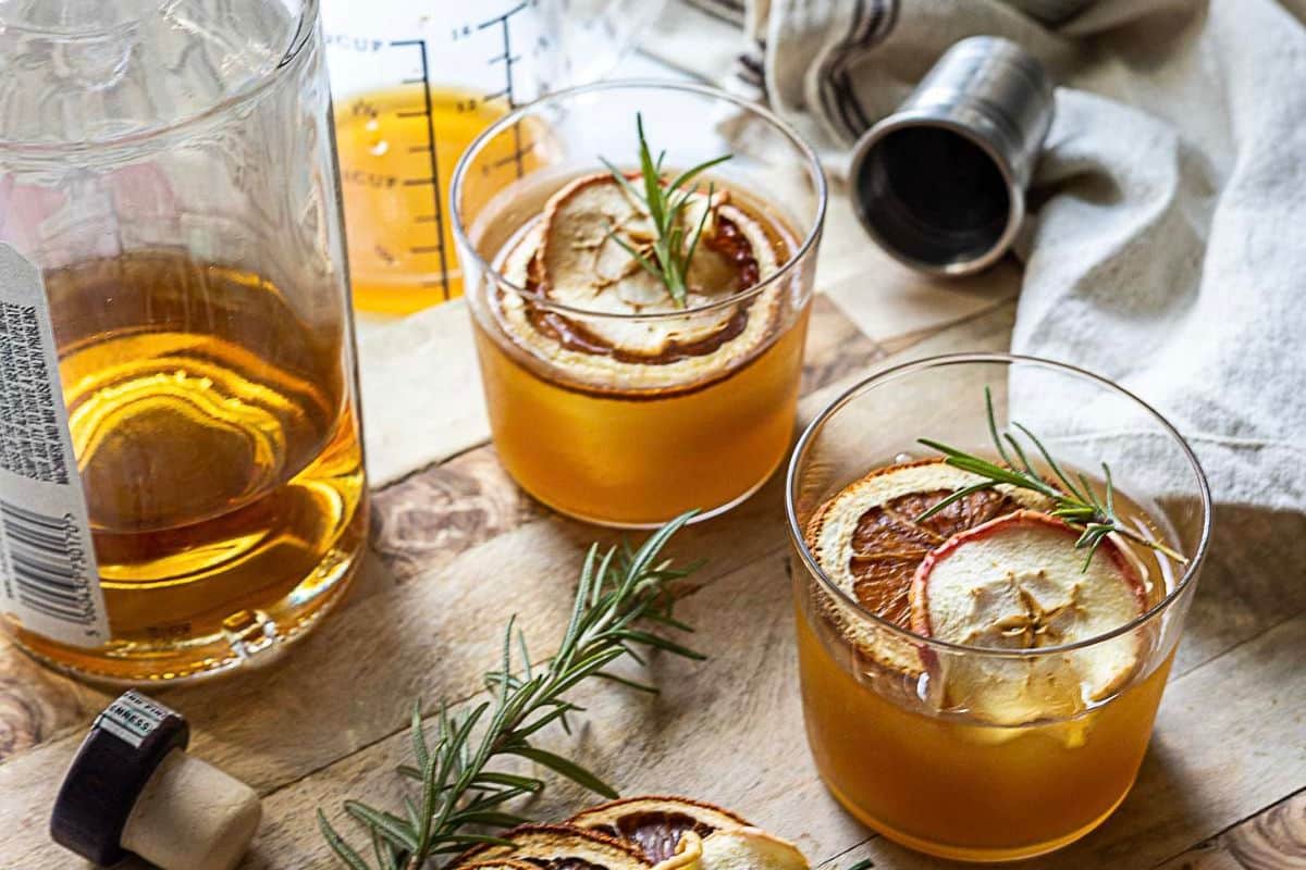 Two glasses of amber apple drinks garnished with dried apple slices and rosemary sit on a wooden surface next to a bottle of liquor, a jigger, and a linen cloth.