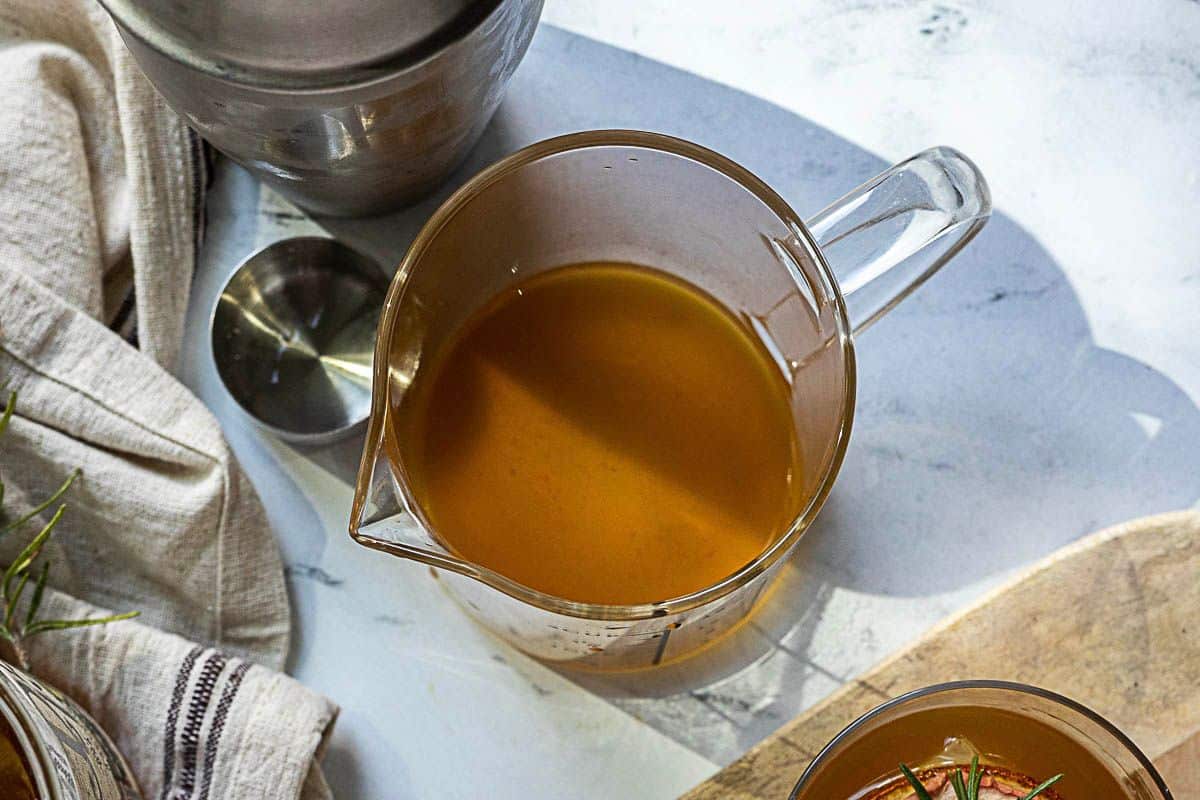 A glass measuring cup filled with brown broth sits on a light-colored countertop next to a metal utensil and a striped cloth, an inviting scene for preparing delicious apple recipes or warming apple drinks.