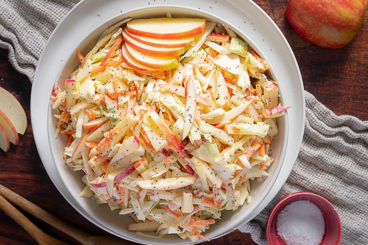 A bowl of apple coleslaw with shredded cabbage, carrots, and apple slices, garnished with black pepper on a wooden table-a refreshing choice for those seeking new apple recipes or light apple snacks.