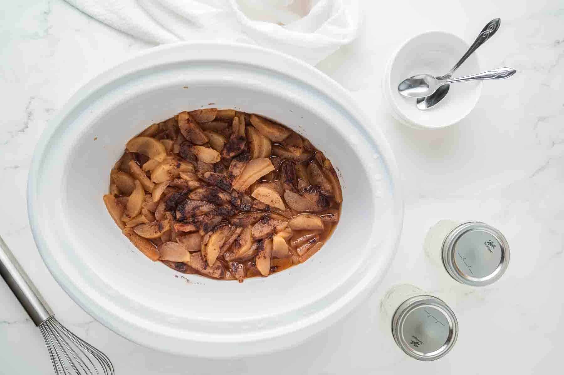 Sliced apples with cinnamon and spices in a white slow cooker, with a whisk, two jars, and a bowl with two spoons nearby on a white countertop.