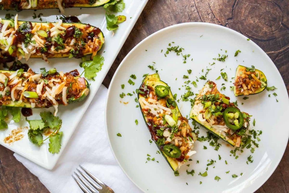 A white plate with stuffed zucchini boats topped with cheese, herbs, and sliced jalapeños sits on a wooden table alongside a fork and a platter of more zucchini-perfect for family dinners or quick dinner recipes.