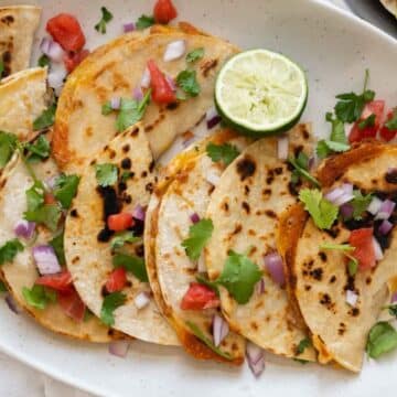 Four folded, grilled tacos topped with chopped onions, tomatoes, and cilantro are arranged on a white oval plate with half a lime—perfect for quick family meals or easy 30 minute dinners.