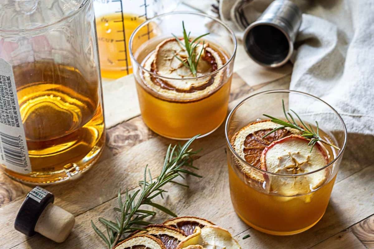 Two glasses of amber-colored Fall cocktails, garnished with dried apple slices and rosemary, rest on a wooden surface beside a whiskey bottle, jigger, and cocktail ingredients-perfect seasonal drinks for autumn gatherings.