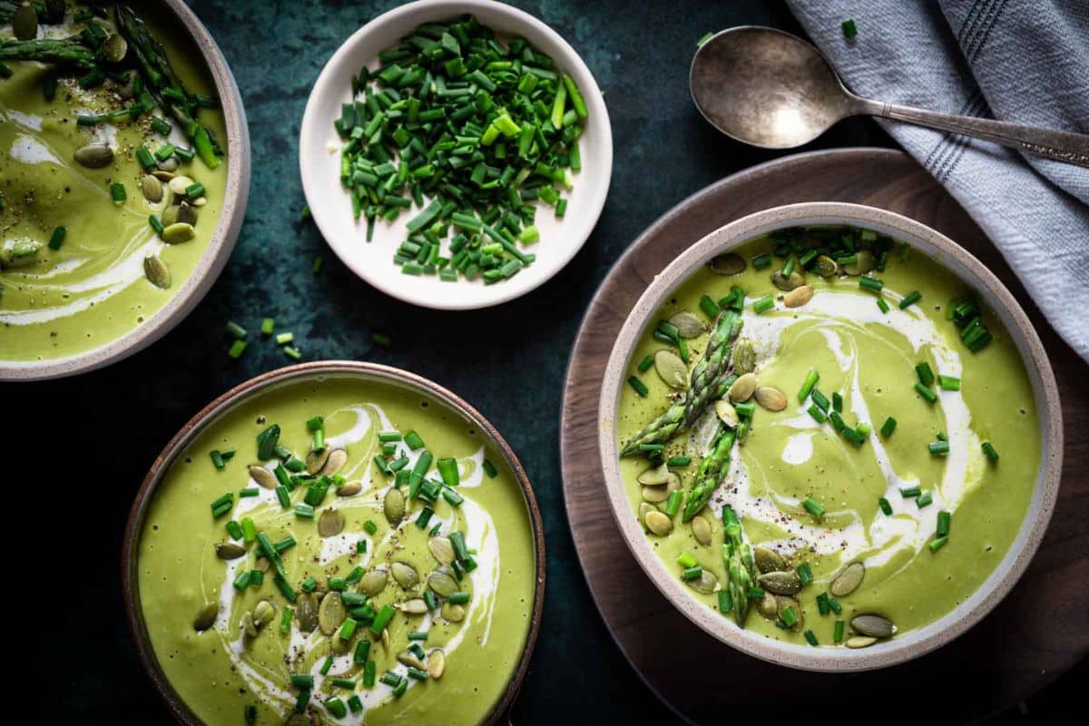 Three bowls of green, creamy soup topped with cream, chopped chives, pepitas, and asparagus sit alongside a small bowl of chives and a spoon on a dark surface-perfect for fans of cozy soups and fall soup recipes.