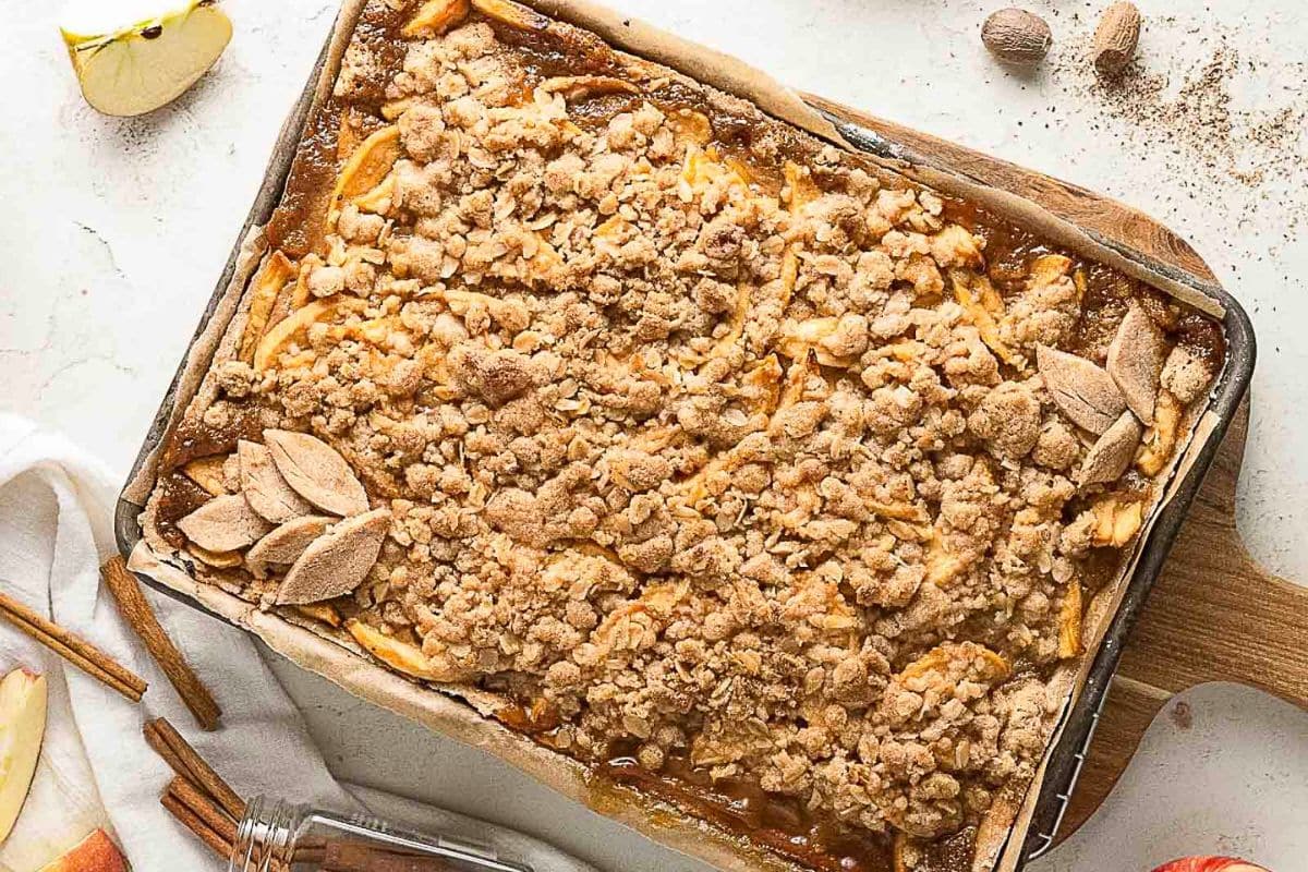 A rectangular baking tray filled with apple crumble bars, topped with crumbly streusel and small leaf-shaped pastry decorations. Sliced apples and cinnamon sticks are nearby, perfect for fans of sweet apple recipes.