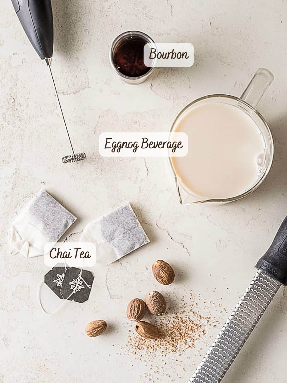 Overhead view of eggnog cocktail ingredients, including bourbon, eggnog beverage, chai tea bags, nutmeg, a grater, a frother, and a small bowl of liquid on a light surface.