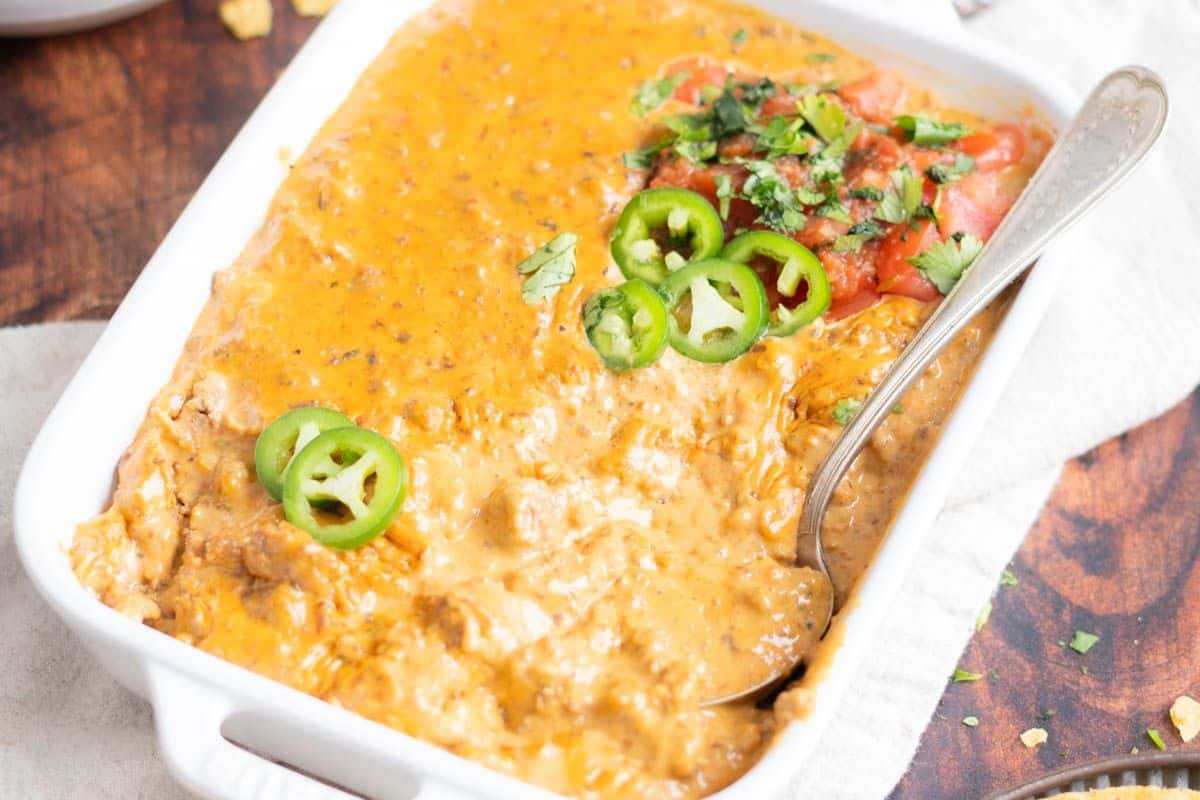 A baking dish filled with cheesy bean dip topped with sliced jalape&ntilde;os, chopped cilantro, and salsa-perfect for Game Day Dips-with a spoon resting inside.