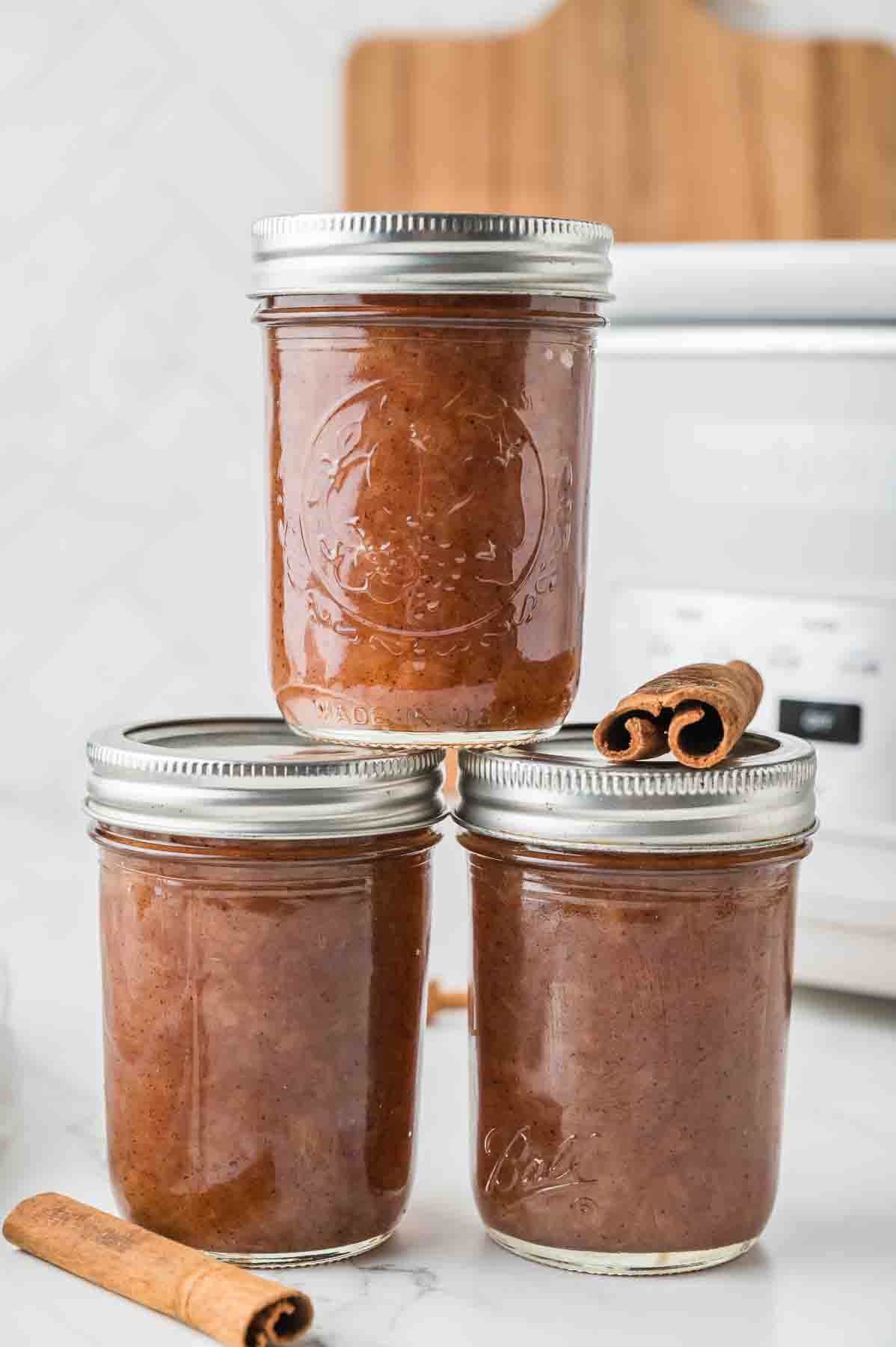Three glass jars filled with apple sauce are stacked on a countertop, accompanied by two cinnamon sticks.