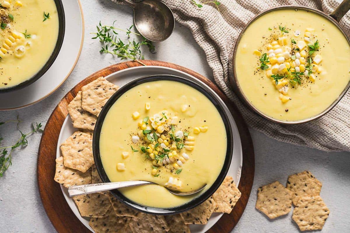 Two bowls of creamy corn soup-perfect for cozy soup nights or fall soup recipes-garnished with corn, herbs, and black pepper, served with crackers on a wooden tray, alongside a cloth napkin and spoons.