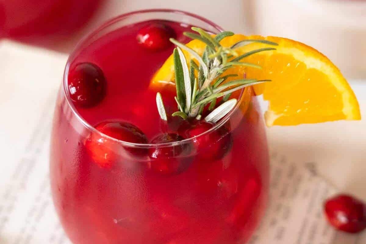 A glass of red cranberry drink garnished with whole cranberries, a sprig of rosemary, and a slice of orange-the perfect choice for autumn drinks and seasonal cocktails.