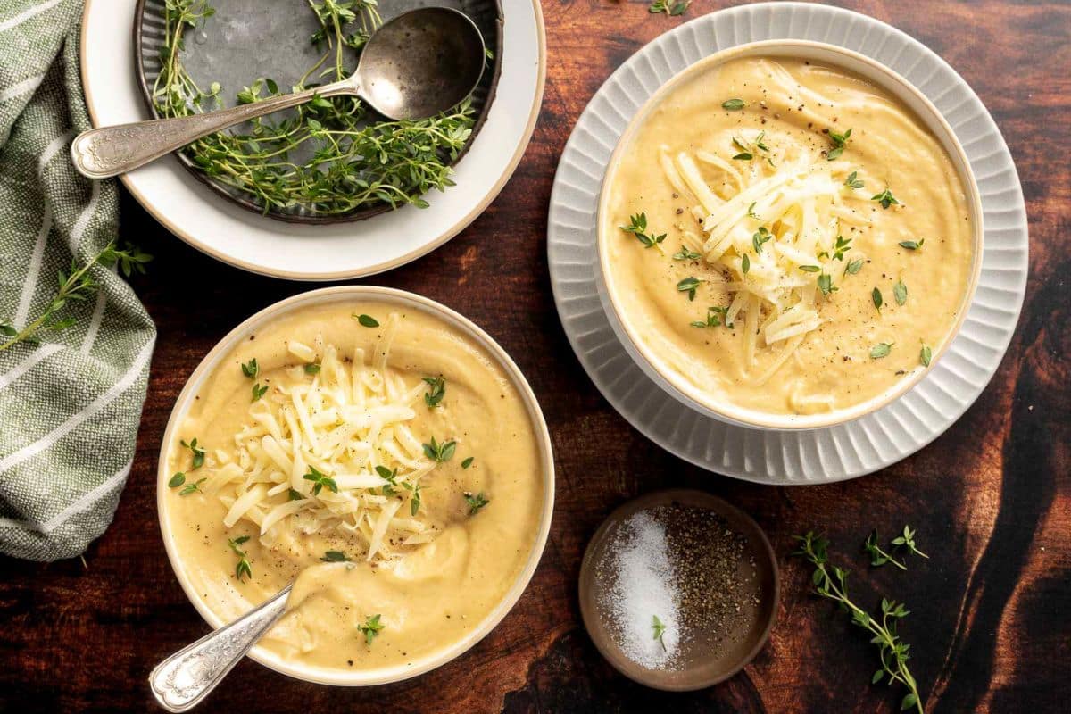 Two bowls of creamy cheese soup, one of our favorite cozy soups, are topped with shredded cheese and fresh thyme. A dish of pepper and salt, plus a plate of thyme and spoons, sit nearby on the wooden table-perfect for fall soup recipes.