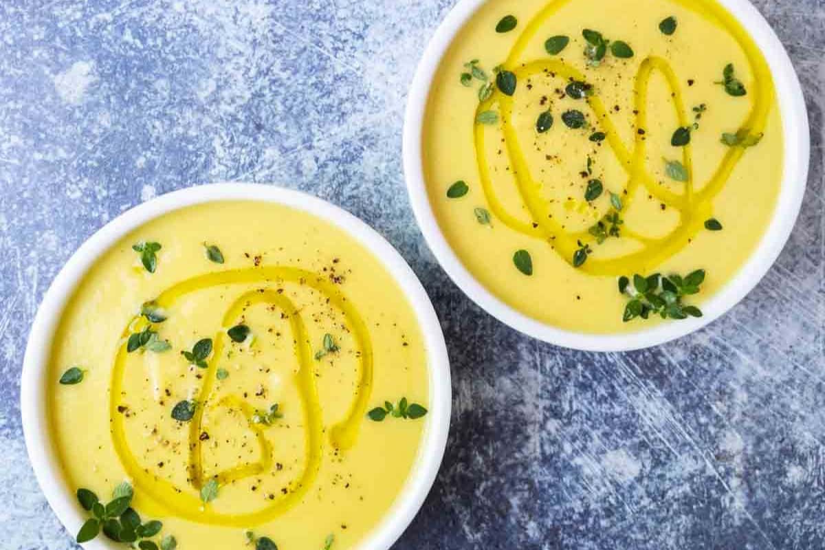 Two bowls of creamy yellow soup, a perfect addition to your favorite fall soup recipes, garnished with fresh herbs, black pepper, and a drizzle of olive oil on a textured gray surface.