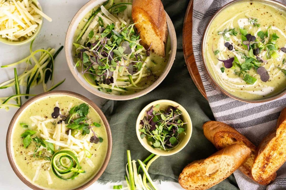 Three bowls of creamy green soup, one of our favorite cozy soups, are topped with microgreens, shredded cheese, and spiralized zucchini, served with toasted bread slices and extra toppings on the side.
