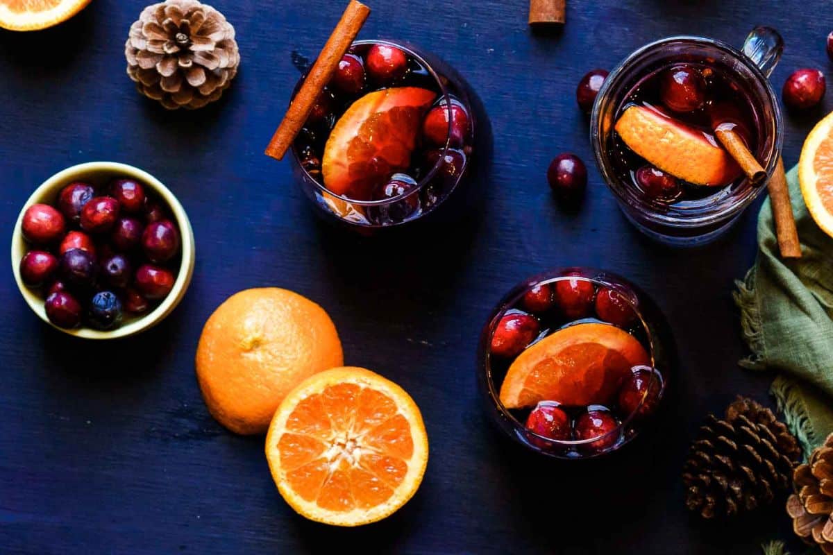 Top view of glasses filled with a dark beverage, orange slices, cranberries, and cinnamon sticks-surrounded by fresh oranges, cranberries, pinecones, and a green cloth-perfect inspo for cozy fall cocktails or festive seasonal cocktails.