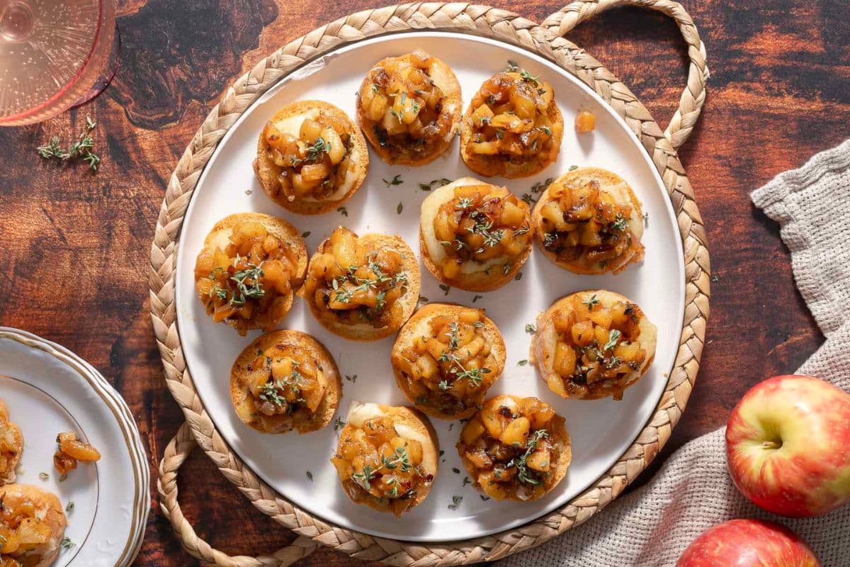 A plate of mini puff pastry tarts topped with a savory apple and herb mixture, placed on a woven placemat with apples nearby on a wooden table-perfect for those seeking creative savory apple recipes.
