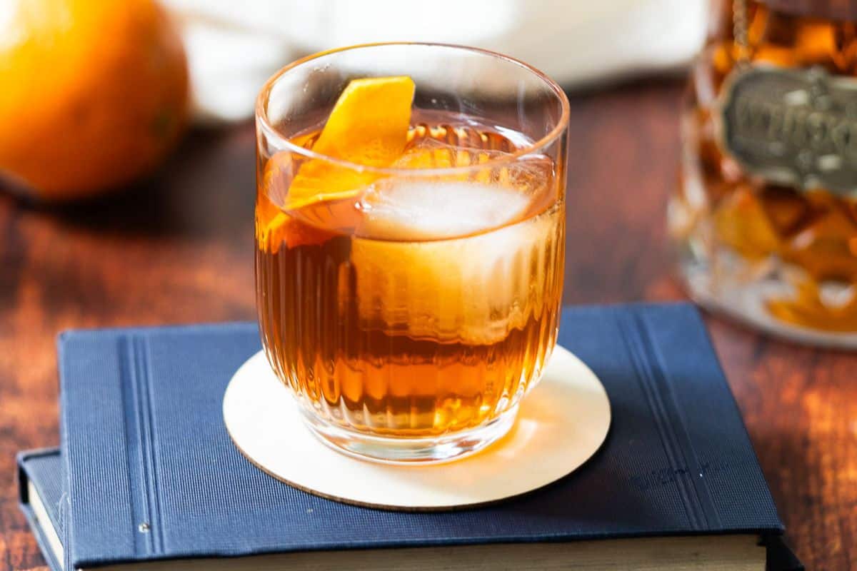 A glass of amber-colored cocktail with ice and an orange peel garnish sits on a coaster atop a closed book, capturing the cozy essence of autumn drinks.