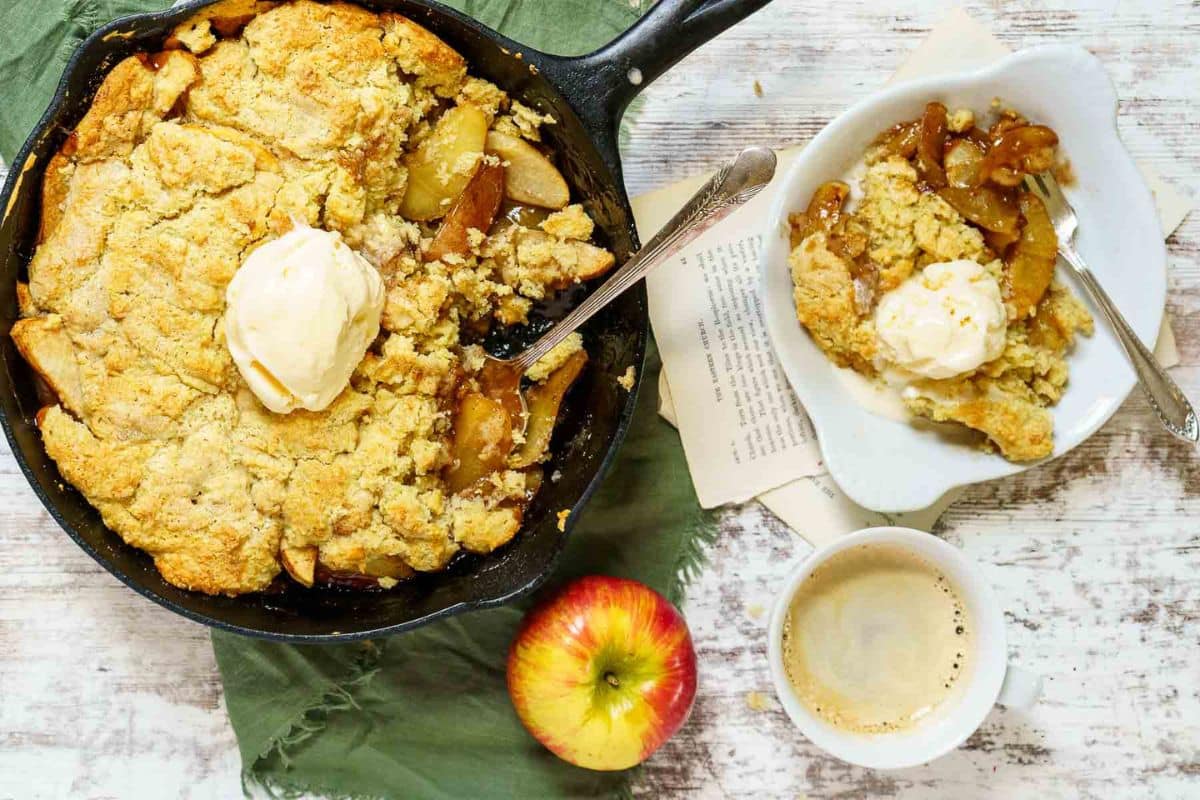 A skillet apple cobbler topped with vanilla ice cream, served with a portion on a plate, an apple, and a cup of coffee on a rustic table-one of the sweetest apple recipes for cozy gatherings.