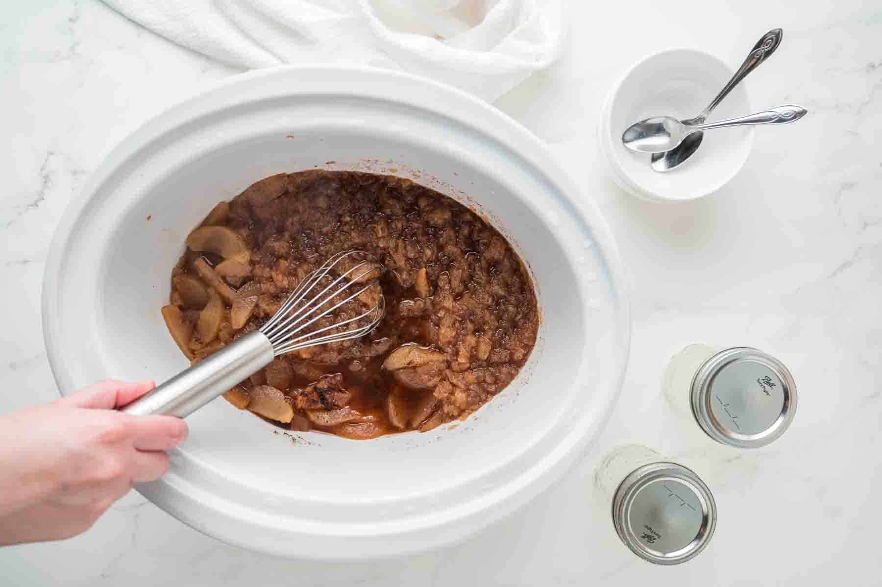 A hand stirs apple mixture with a whisk in a white slow cooker, with two empty jars, a bowl, and spoons nearby on a white countertop.