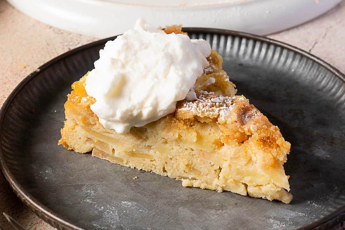 A slice of apple cake topped with a dollop of whipped cream on a dark plate-perfect for those who love apple snacks and homemade Apple recipes.