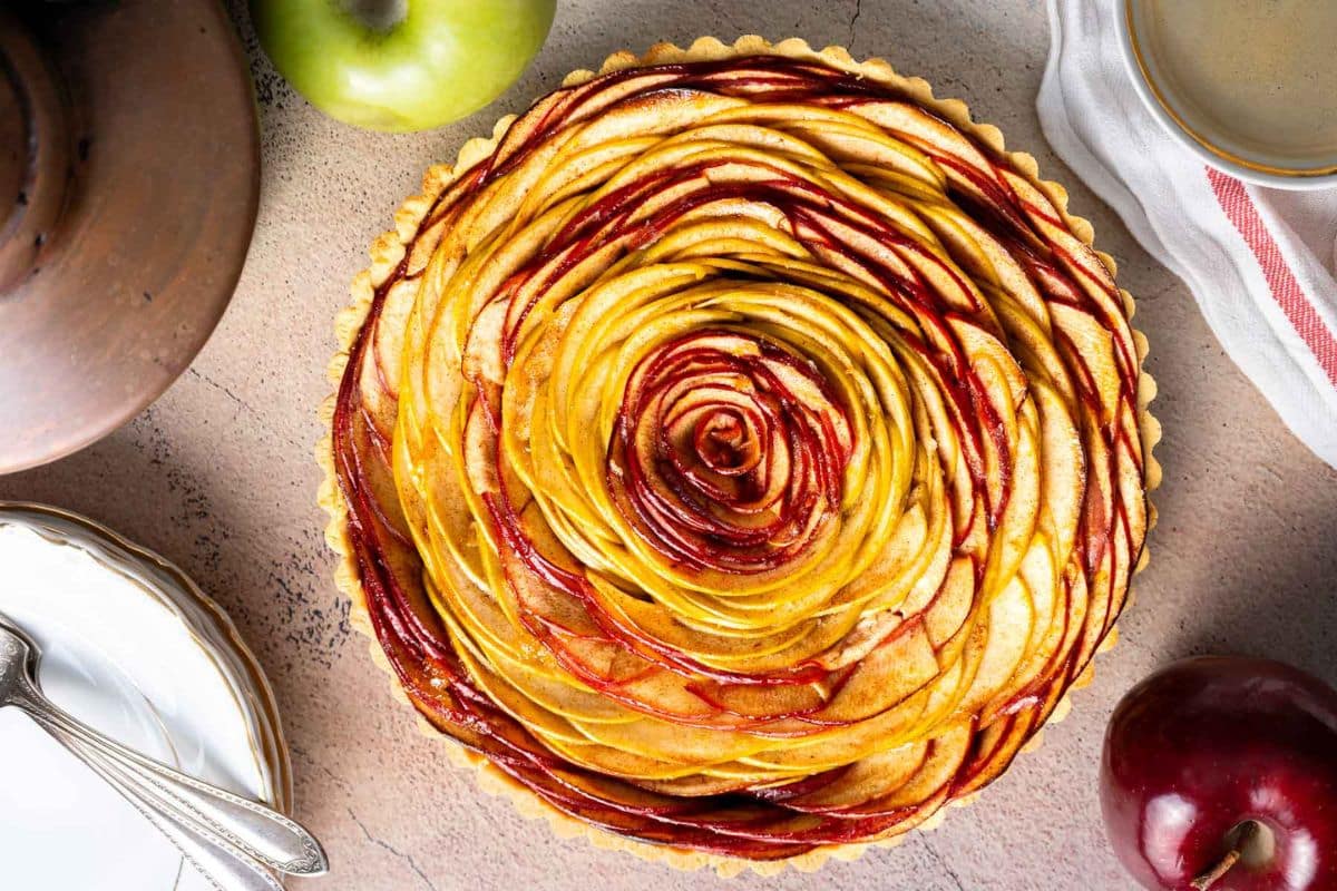 A baked apple tart with thinly sliced apples arranged in a rose pattern, surrounded by a green apple, a red apple, a plate, and a cup-perfect inspiration for your next sweet apple ideas.