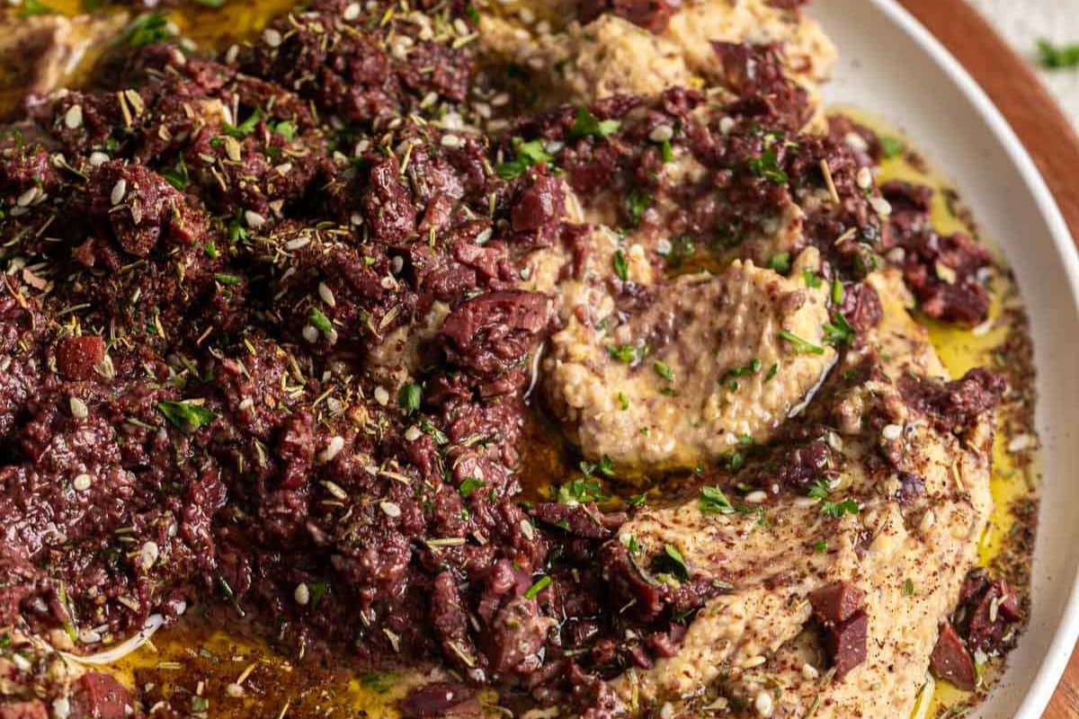 A close-up of hummus topped with chopped olives, herbs, olive oil, and spices, served on a white plate-a perfect choice for Game Day Dips or Touchdown Snacks.