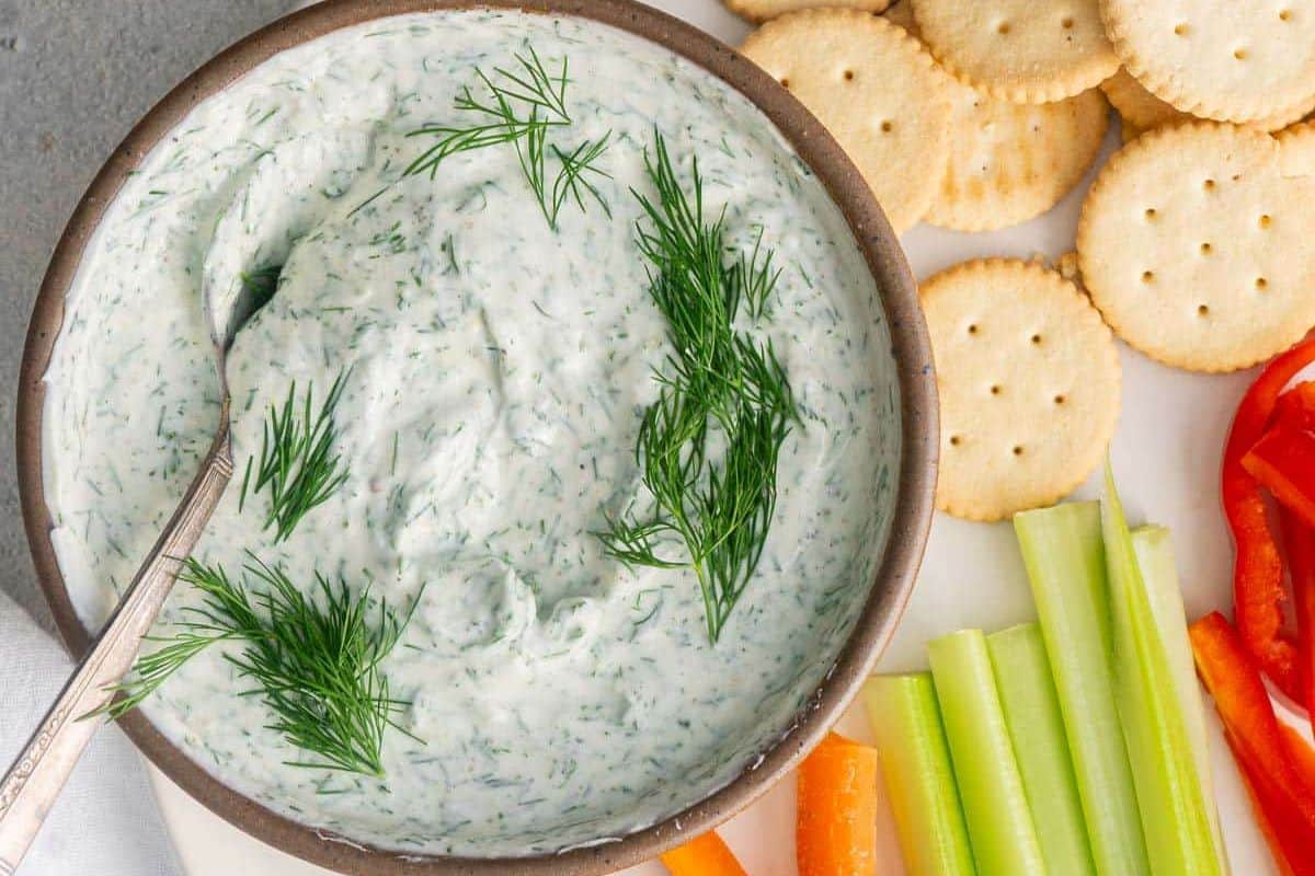A bowl of creamy herb dip garnished with dill, served with round crackers, celery sticks, carrot sticks, and red bell pepper slices-perfect for your lineup of Game Day Dips.