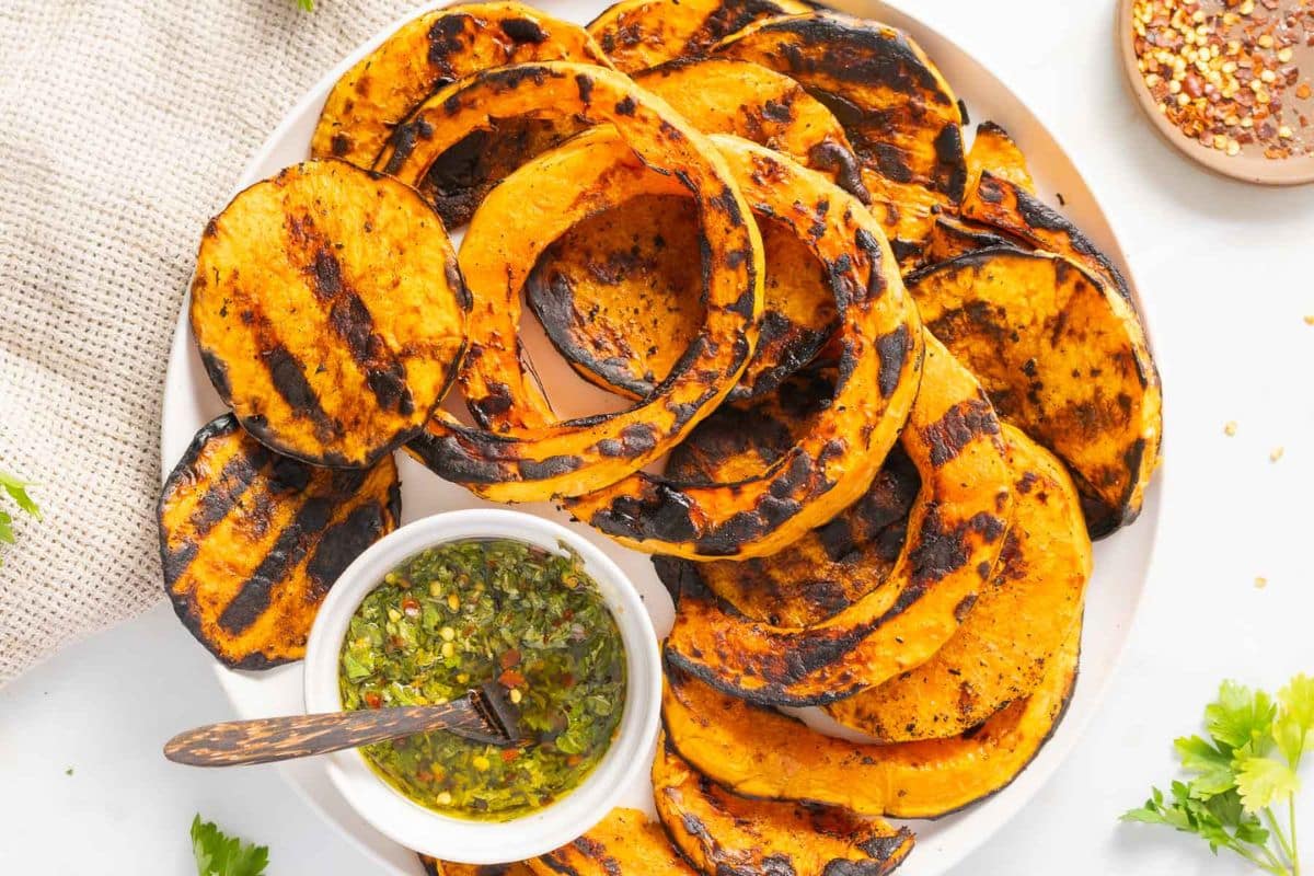 A plate of grilled butternut squash slices with visible char marks, served with a small bowl of green herb sauce and a spoon—perfect for those seeking easy squash recipes.